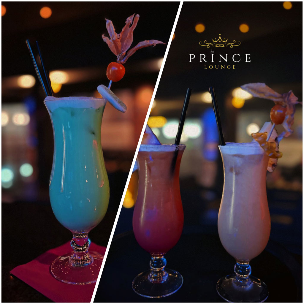 Zwei bunte Cocktails mit Früchte-Deko auf einem Tisch bei Prince Lounge, schwaches Licht.